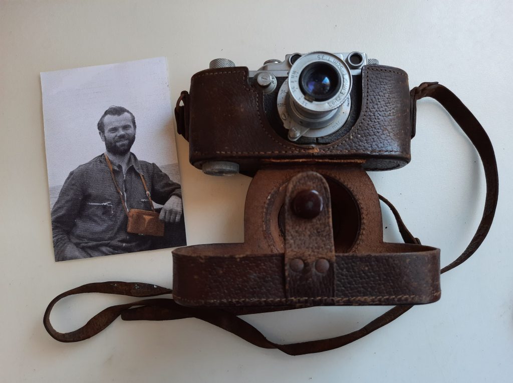 An antique camera with a leather case, positioned next to a black and white photograph of a smiling man wearing a camera around his neck.