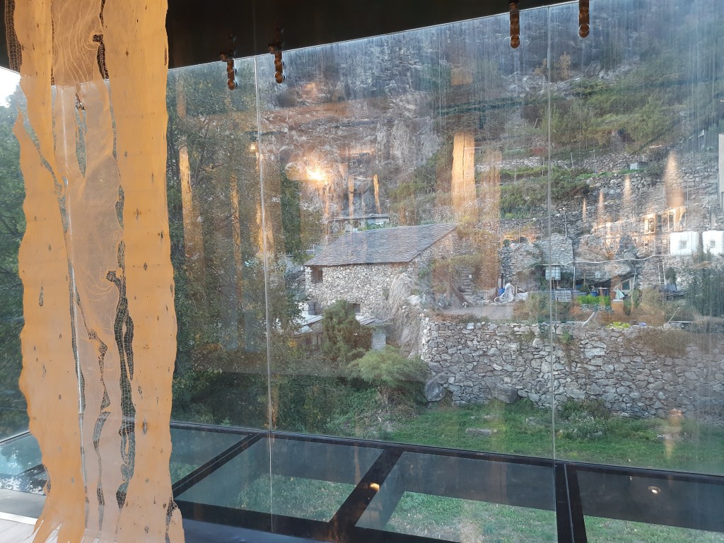 View from an interior space showcasing a large glass window overlooking a rustic stone house and greenery, with cascading decorative elements in the foreground.